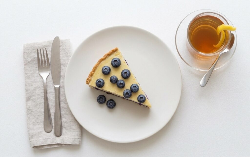 A slice of vegan cheesecake with blueberries on top and a black tea with lemon on the side