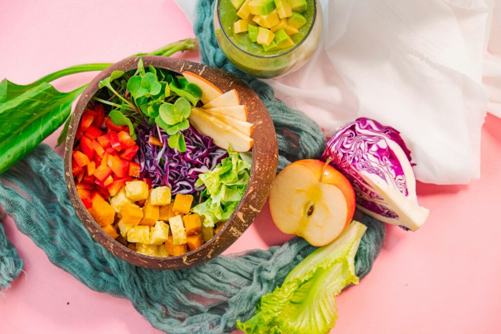 Bowl of fruit and veggies with a smoothie and fruit on the side on a pink background