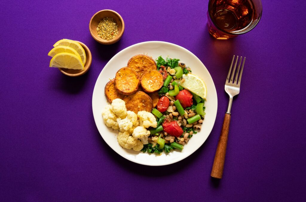 Bowl of veggies and greens with a side of oats and fruit and a drink on a purple background